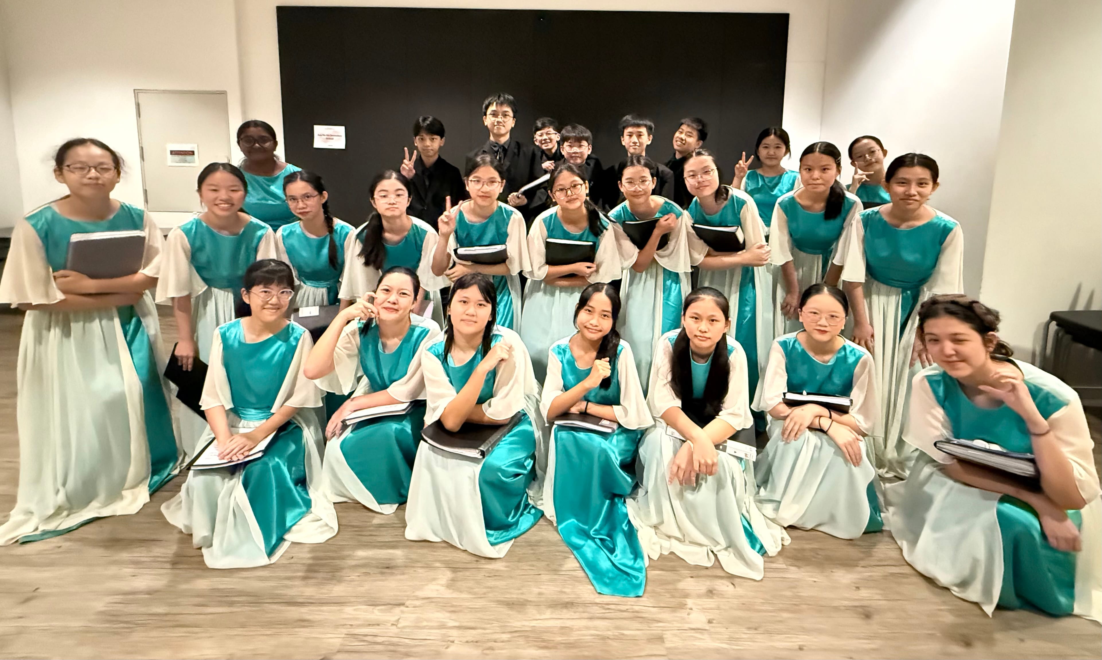 Conductor leads a choir on risers; singers in teal-and-white robes and black suits, pianist at keyboard.